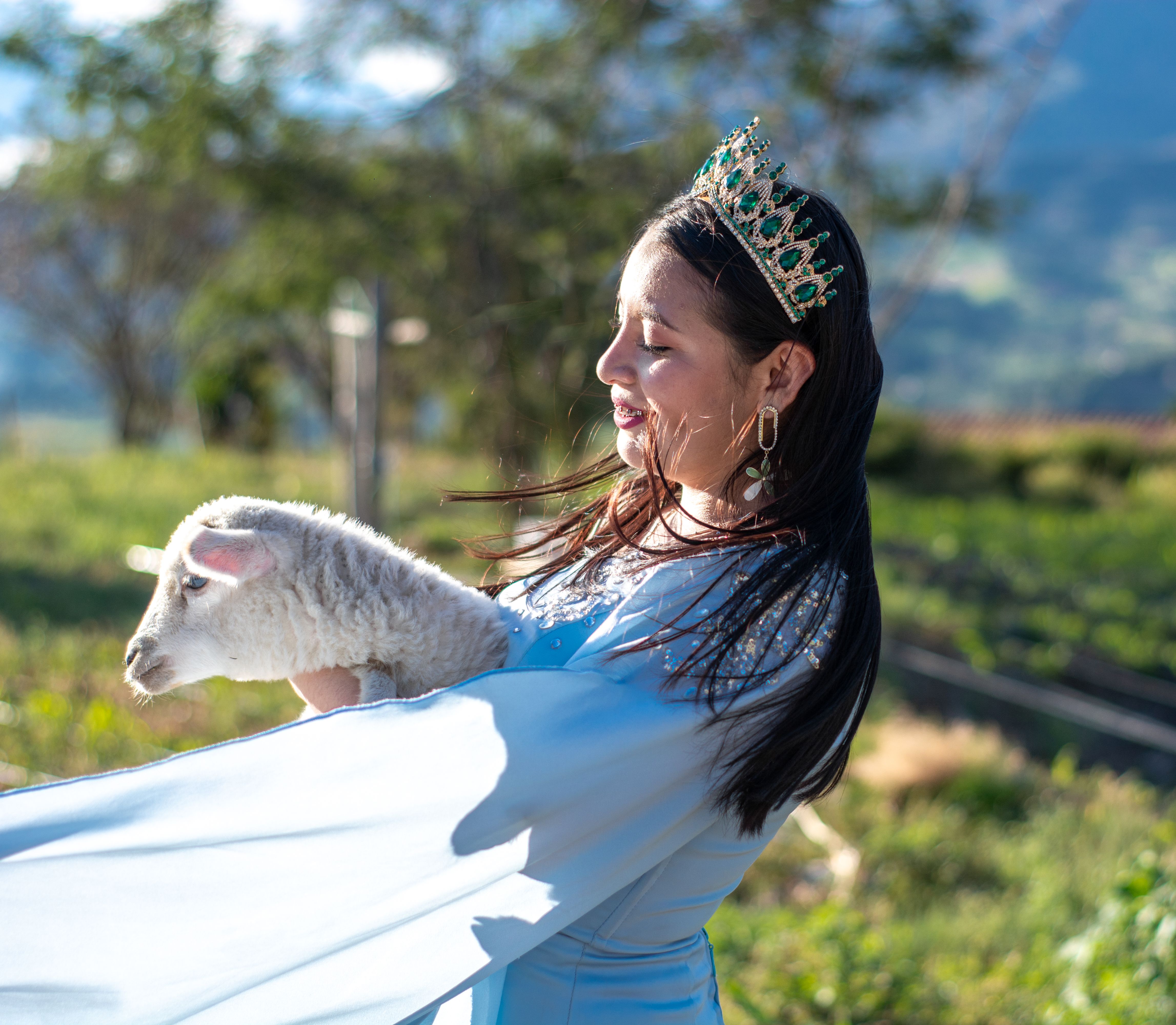A young woman wearing a tiara embraces a lamb in a sunny outdoor setting, showcasing warmth and joy.