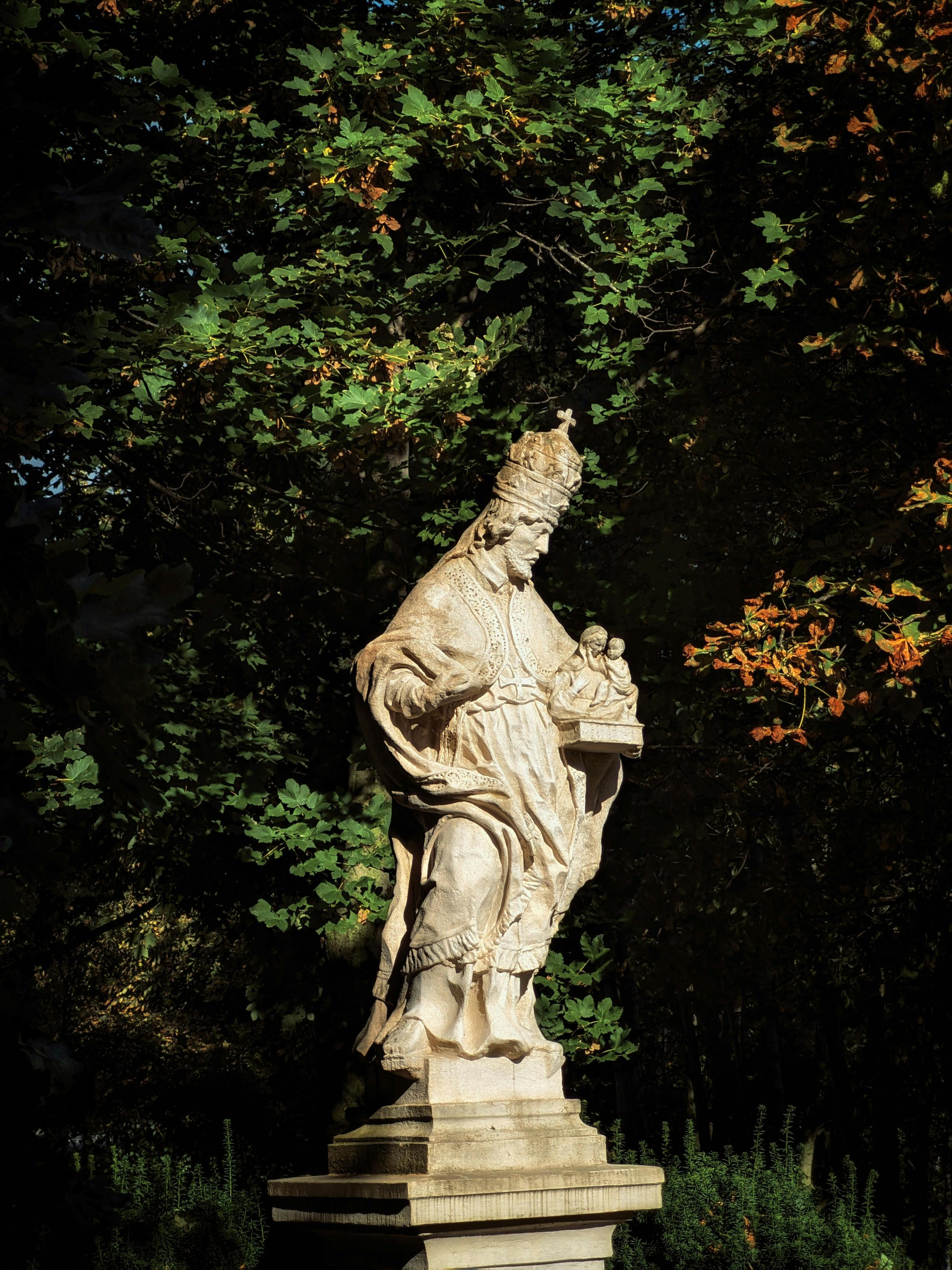 Dramatic stone statue of a religious figure surrounded by lush tree foliage in sunlight.