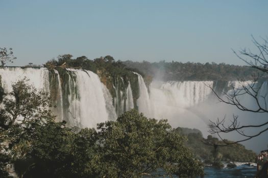 Breathtaking view of foamy waterfall with pond surrounded by mounts and trees under blue sky in daytime