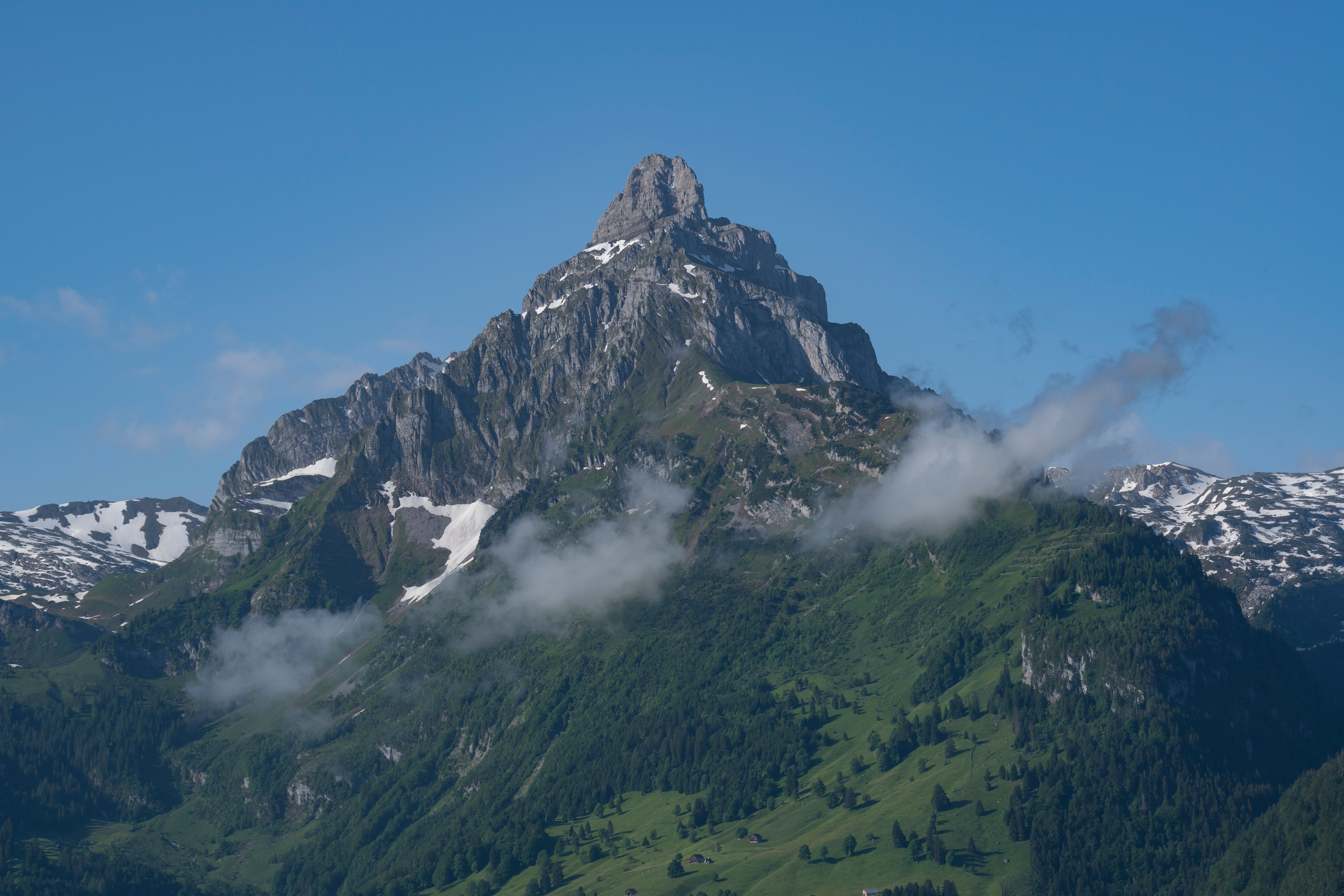 A breathtaking view of a rugged mountain peak surrounded by lush greenery and snow-capped areas under a clear blue sky.