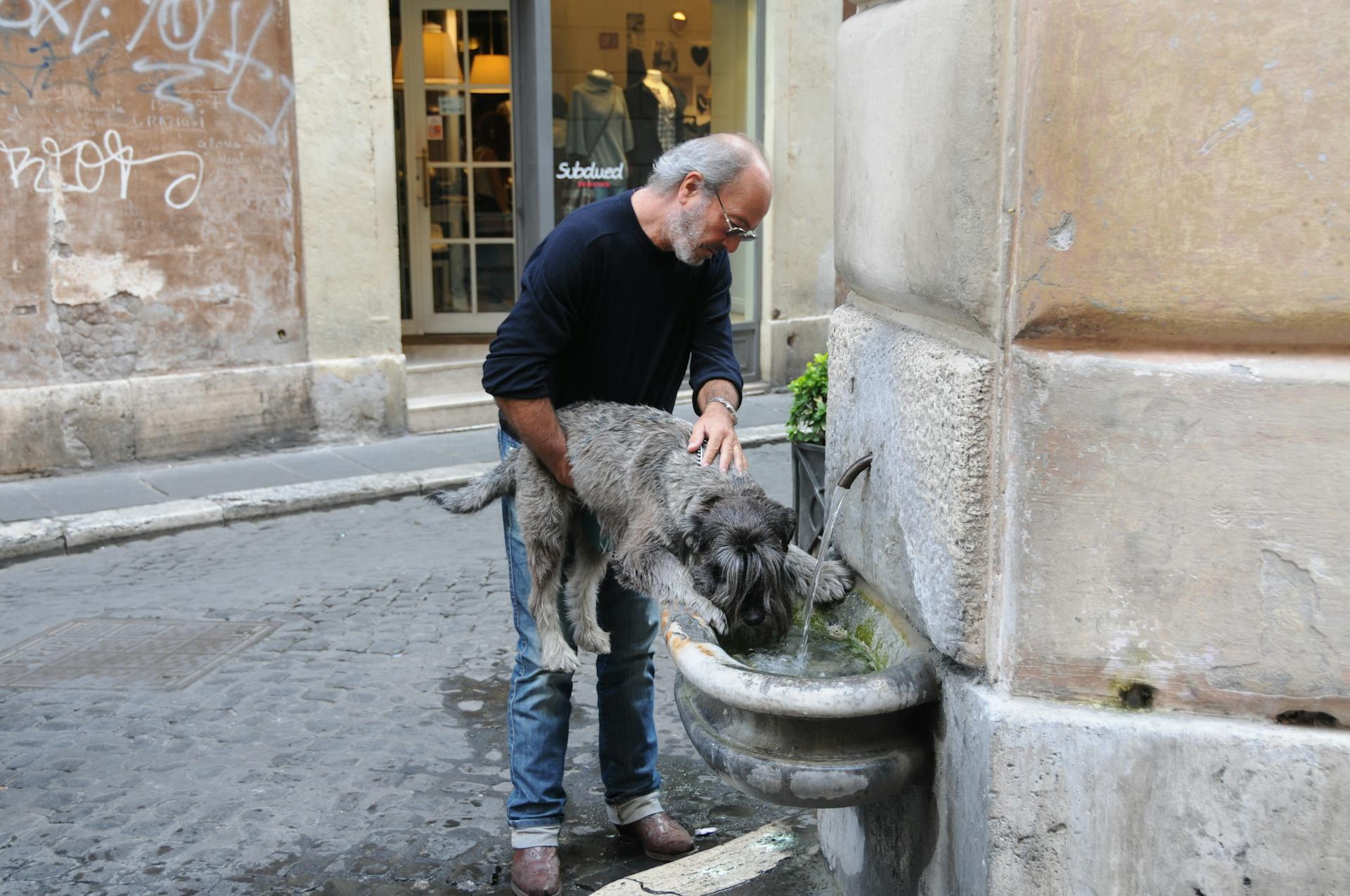 A man assists his dog drinking water from a street fountain in an urban setting.