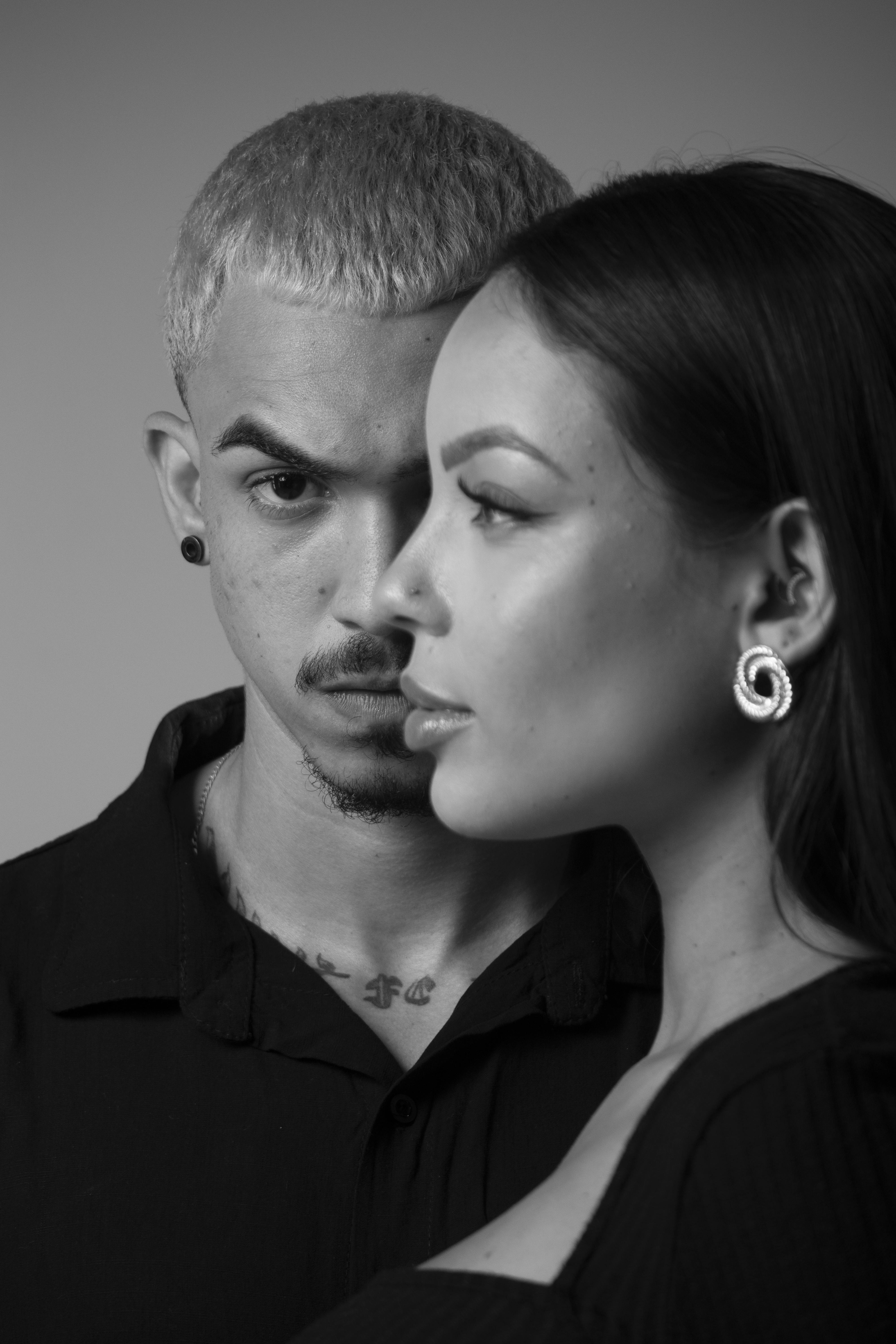 Black and white portrait of two adults with earrings and tattoos, facing forward.