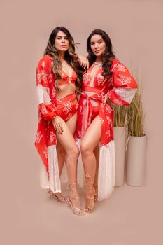 Two women in stylish red beachwear and sandals posing indoors with a neutral background.