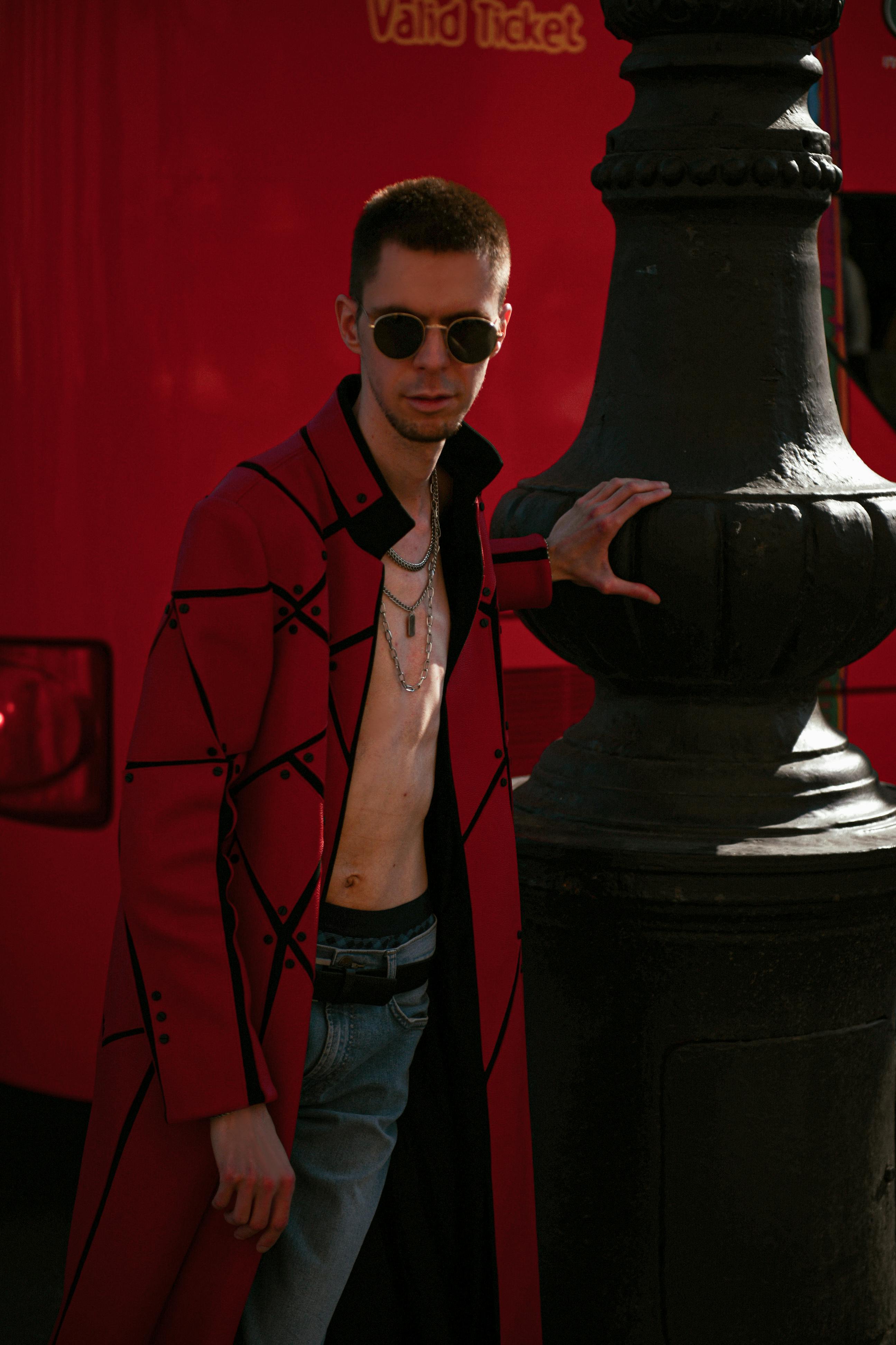 Stylish young man in red coat and sunglasses poses by urban lamp post.