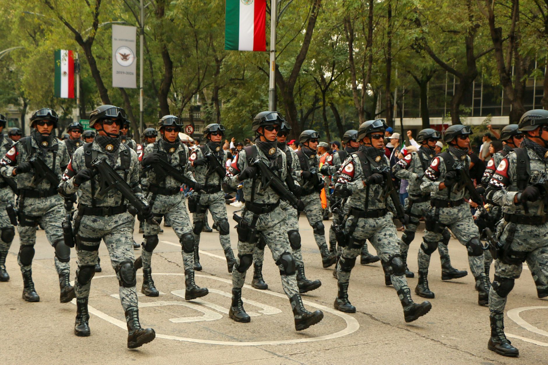 Armed military personnel march in formation during a parade with national flags displayed.