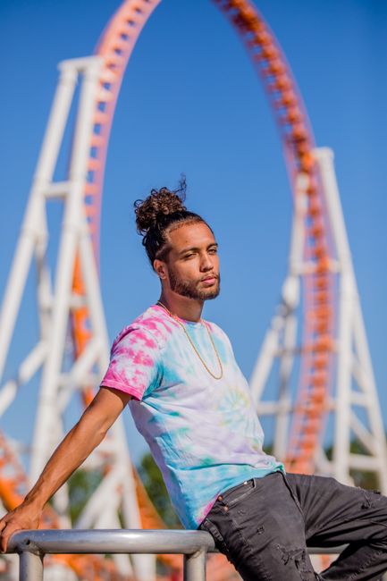 Man in tie-dye shirt relaxing in front of a roller coaster at a sunny amusement park.