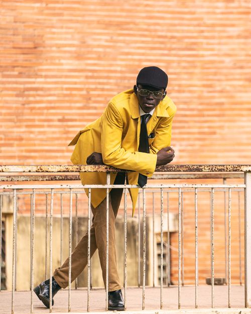 Free Man in yellow coat leaning on railing against brick wall, showcasing urban fashion and style. Stock Photo