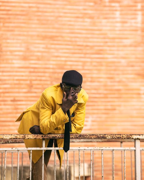Free A fashionable man in a bright yellow suit leans on a rustic balcony, exuding confidence. Stock Photo