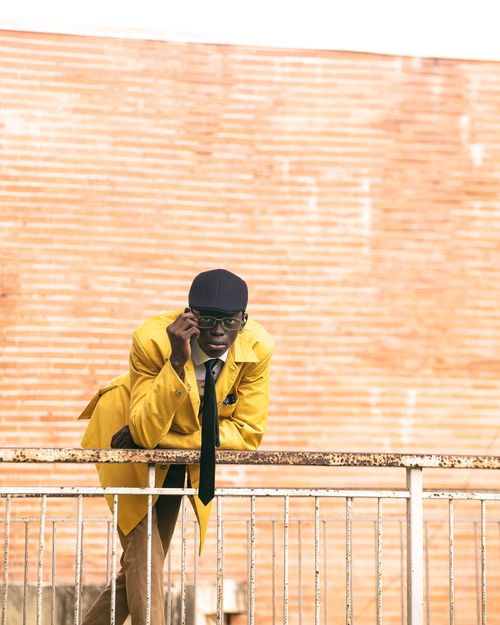 Free A fashionable adult man in a yellow coat leans on a railing against a brick wall backdrop. Trendy urban style. Stock Photo