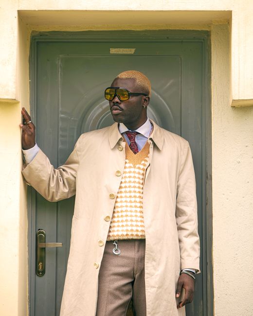 Fashionable young man wearing a beige coat and sunglasses standing at a door frame.