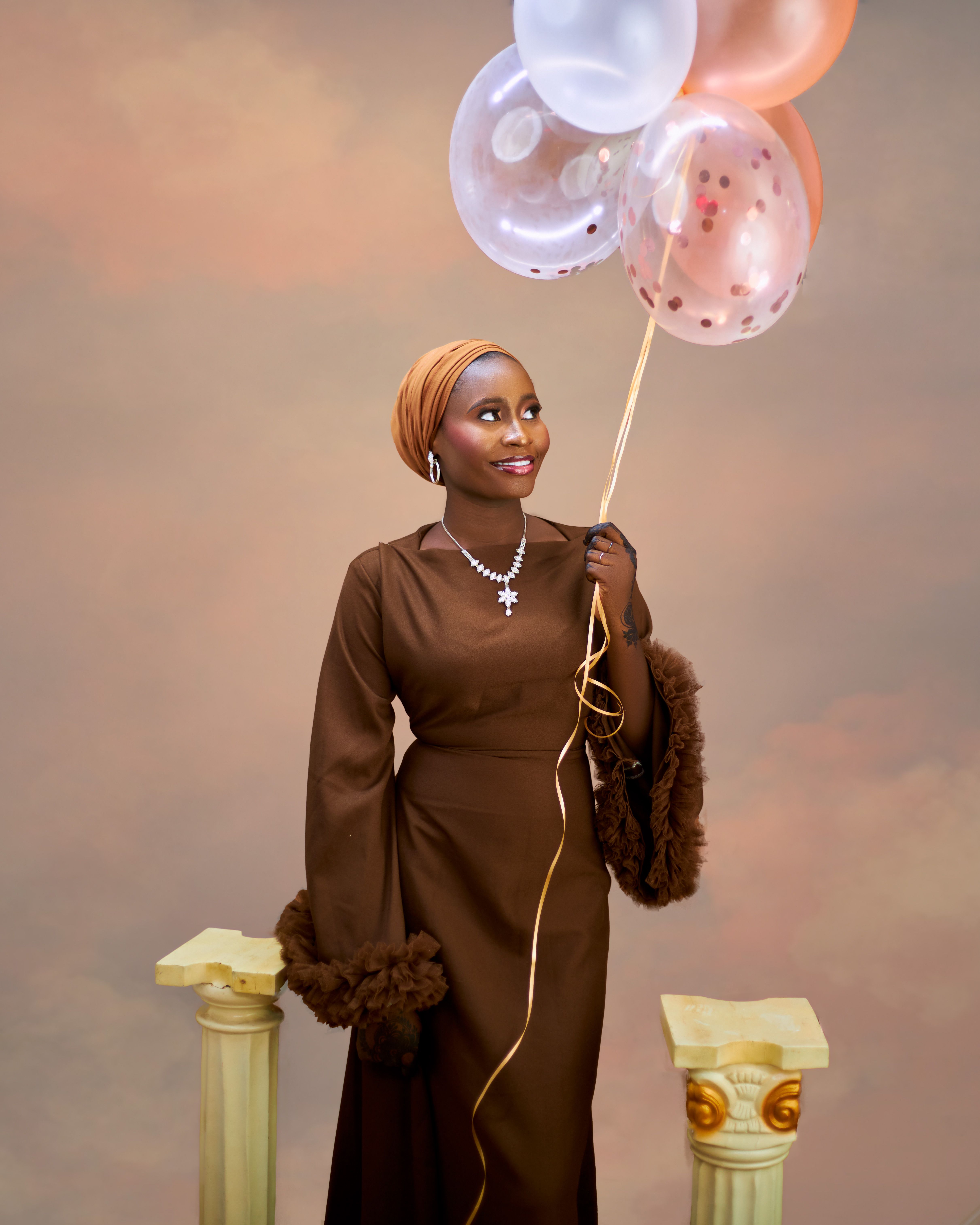 A woman in a brown dress holds colorful balloons against a warm studio backdrop.