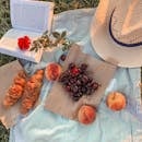 一场风景如画的夏季野餐上，有羊角面包、樱桃、桃子、帽子，以及一条条纹毯子上摊开的一本书