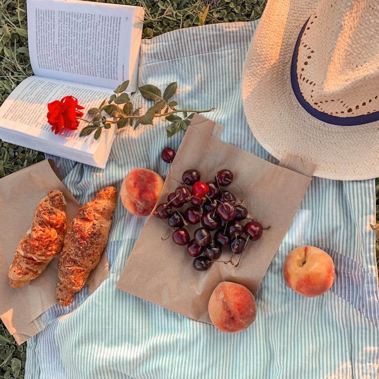 Free A picturesque summer picnic featuring croissants, cherries, peaches, a hat, and an open book on a striped blanket. Stock Photo