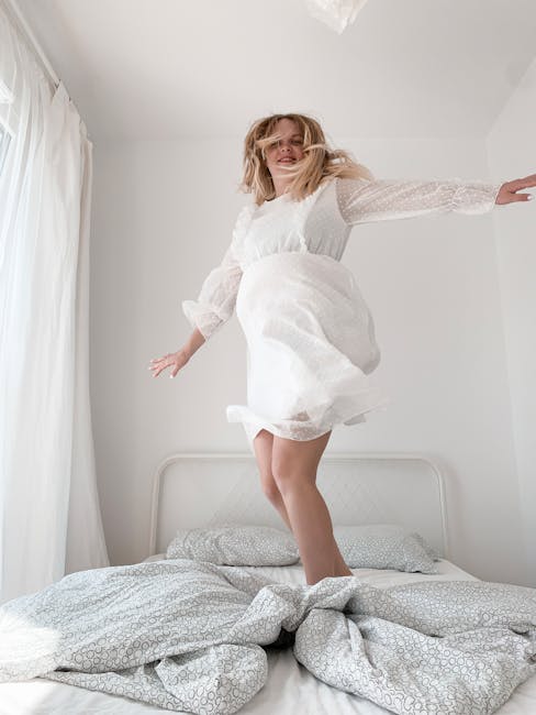 Carefree woman in white dress jumping on bed, expressing joy and happiness.