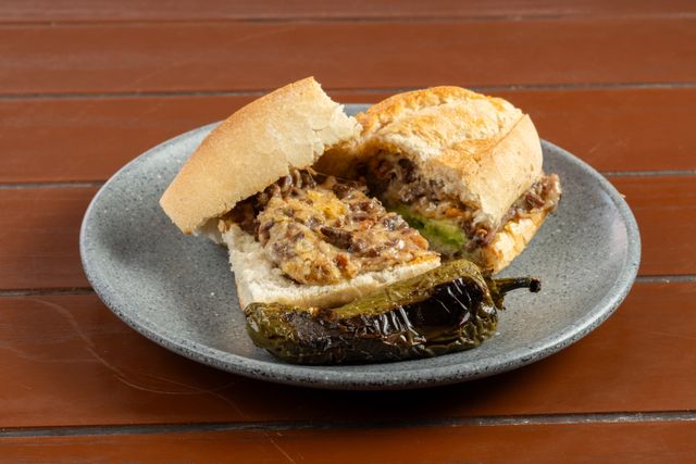 Close-up of a savory cheese and beef sandwich with a side of grilled pepper on rustic wooden table.
