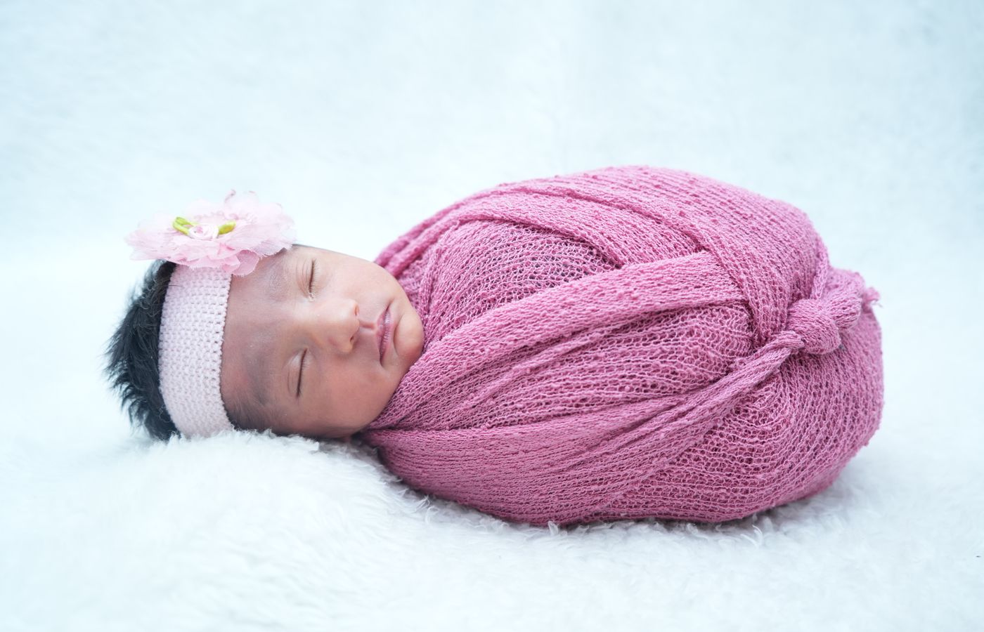 A peaceful newborn baby girl wrapped in a pink knitted blanket with a floral headband.