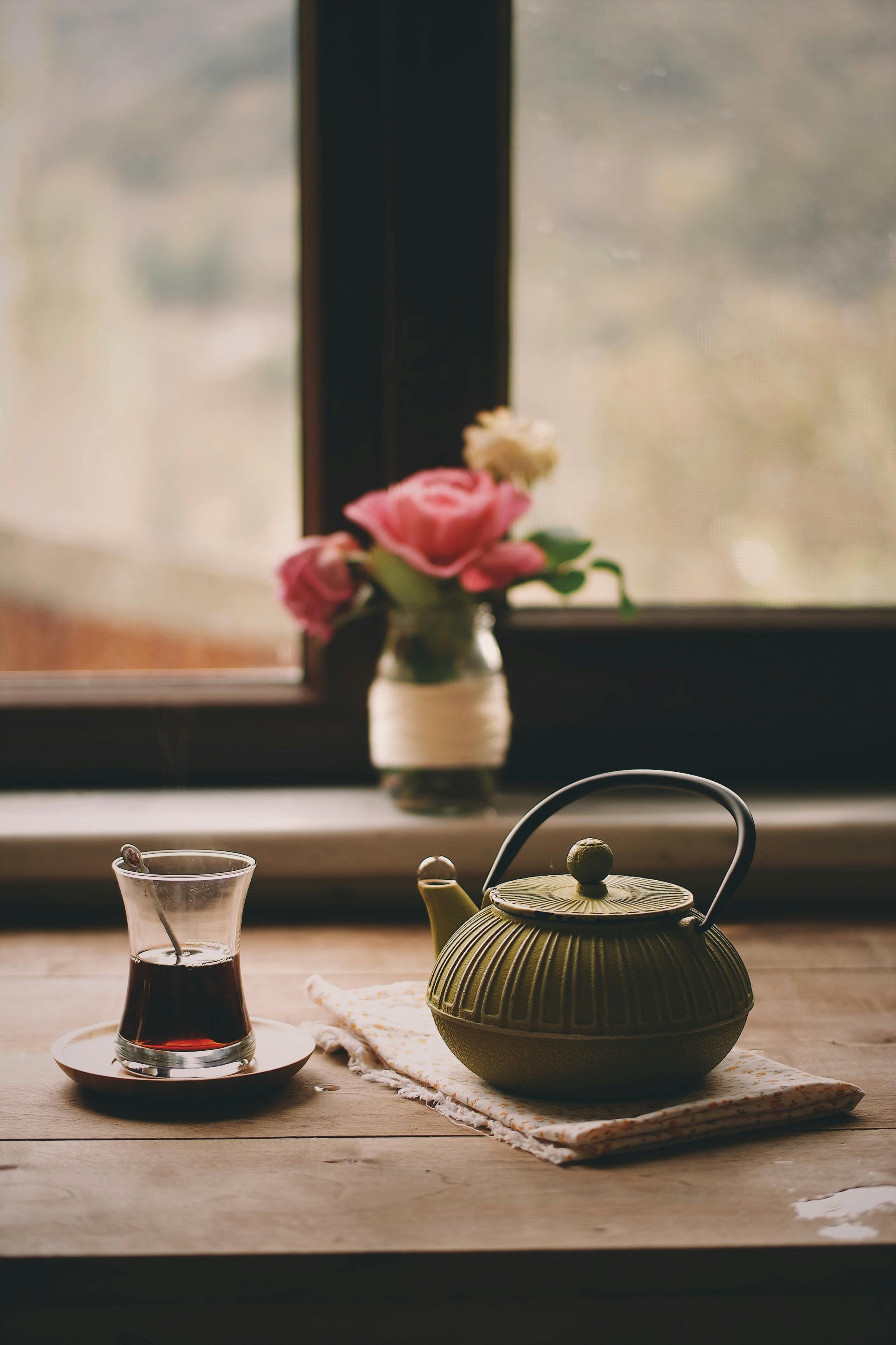 A teapot and glass of tea on a wooden table by a window, with a pink rose bouquet.