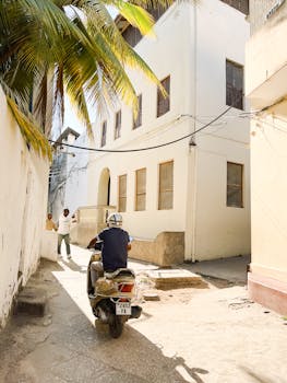 Man riding scooter through sunlit alley in Stone Town, Zanzibar. Vibrant urban scene.