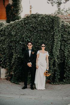 A fashionable bride and groom pose outdoors against a lush ivy backdrop.