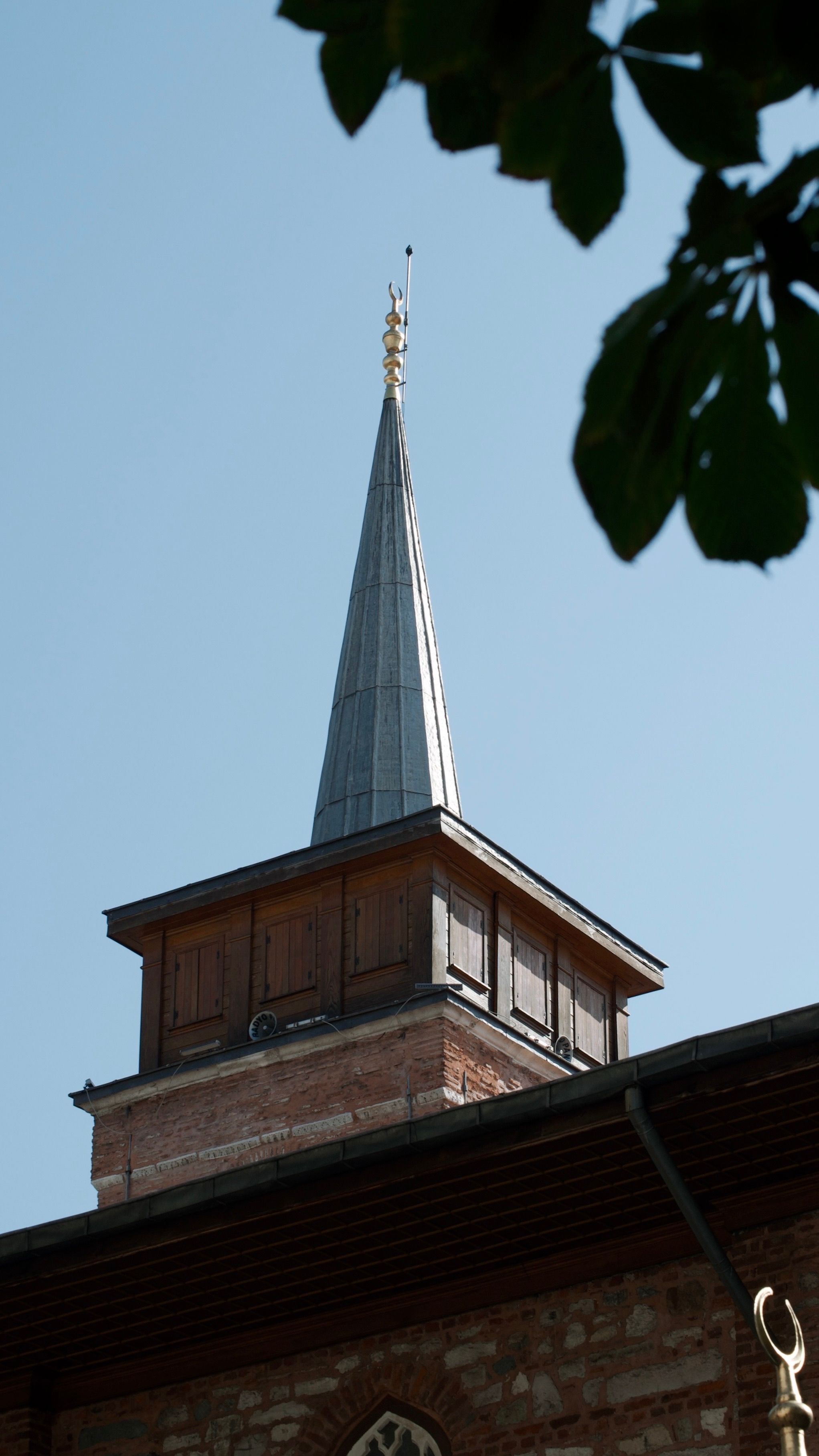 Brick building with a pointed steeple against a clear blue sky, under leafy branches.