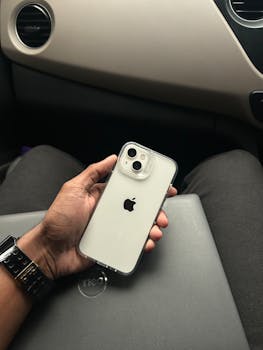 Hand holding a smartphone inside a car on a neutral background.