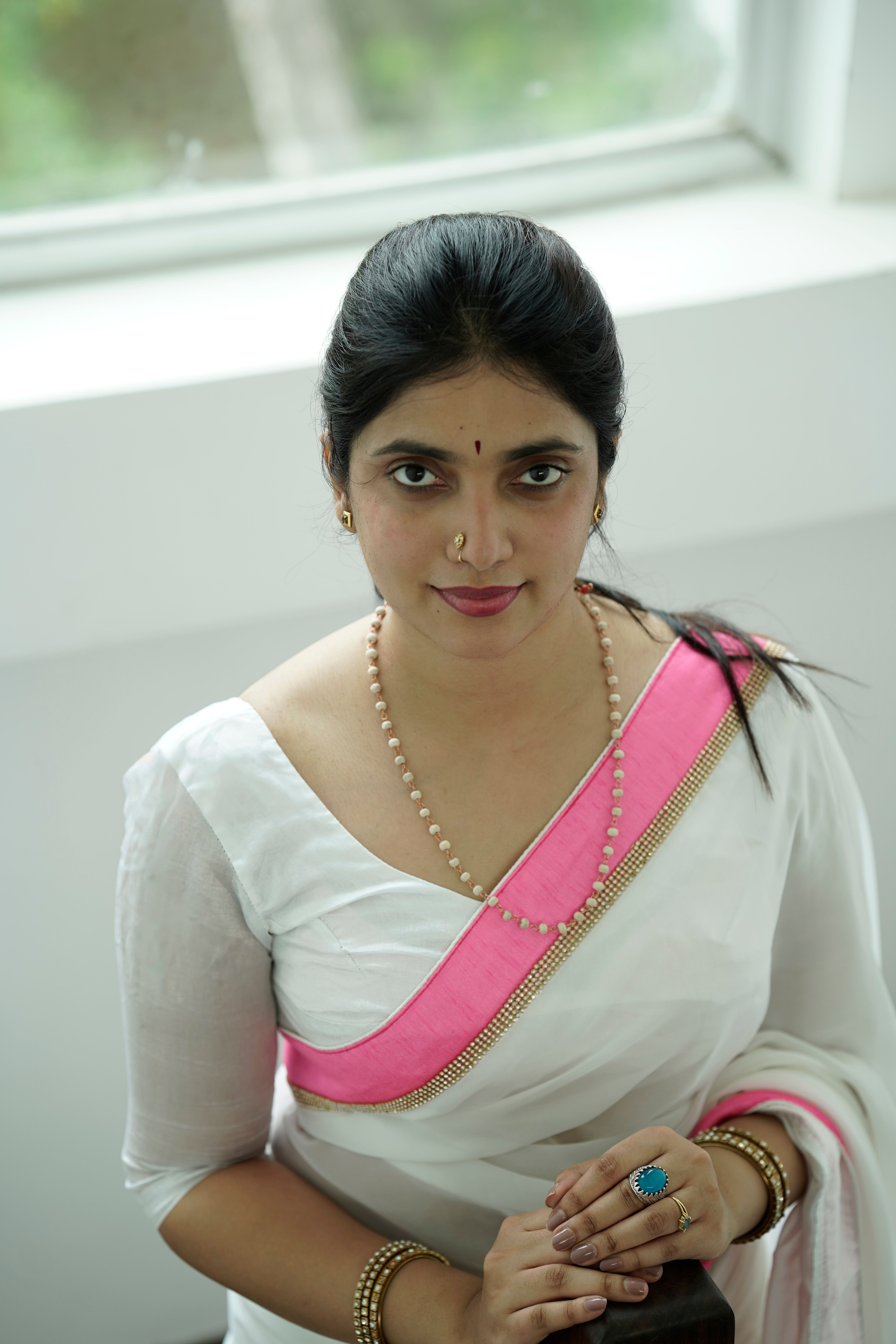 Portrait of a woman in a white saree with pink trim and traditional jewelry.