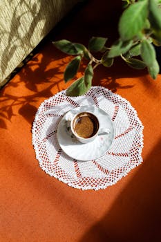 Top view of a coffee cup on a crochet doily with a plant shadow, highlighting simplicity and elegance.