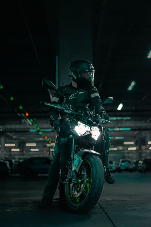 Free Motorcyclist in protective gear sits on parked bike, illuminated in eerie parking garage. Stock Photo