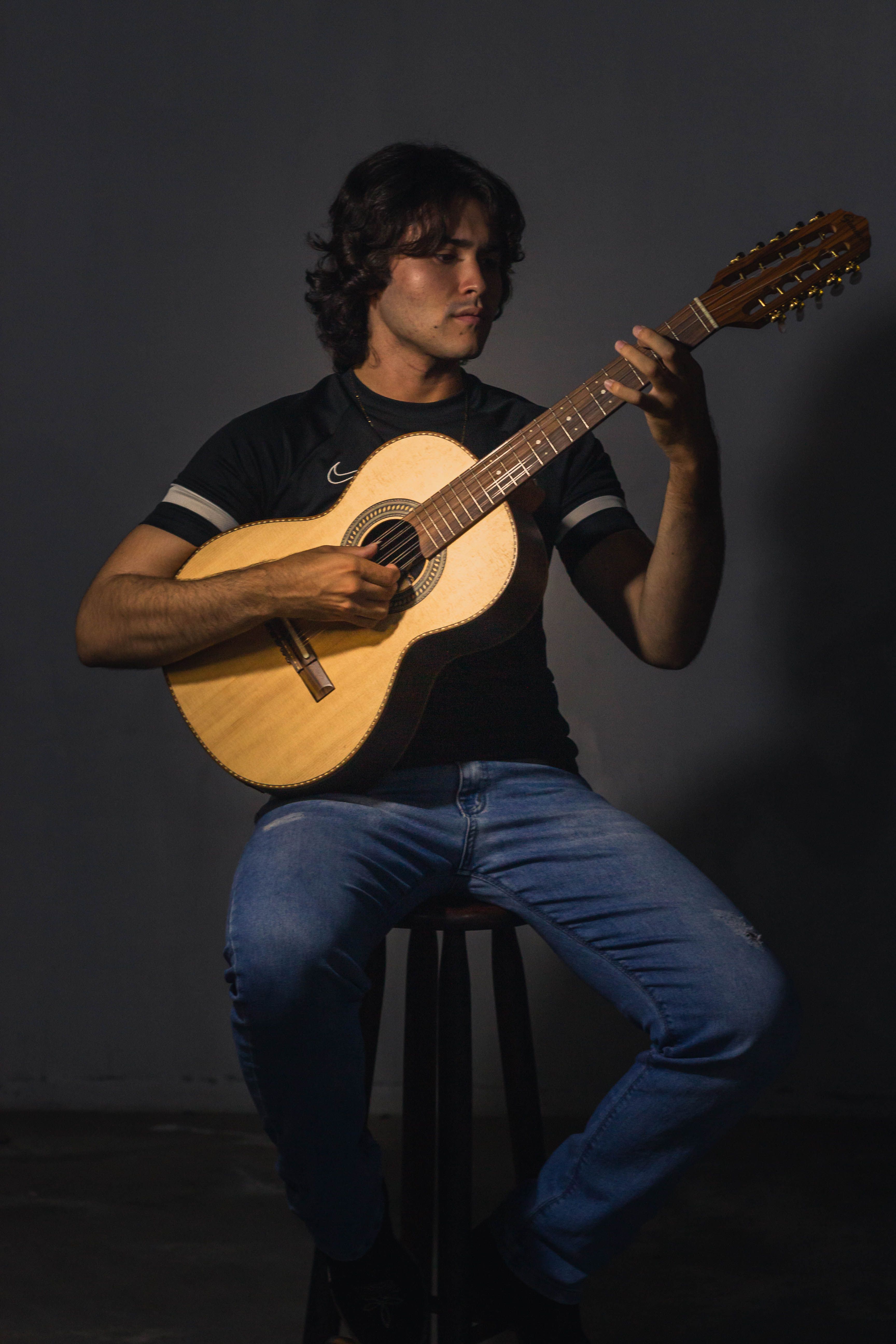 A young man seated on a stool playing an acoustic guitar in a dimly lit room.
