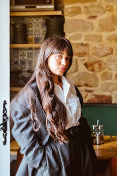Young woman in elegant black coat and white shirt posing indoors with rustic background.