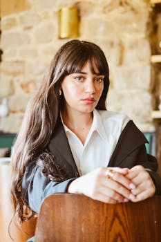 Woman with long hair in a white shirt and black jacket sitting indoors, exuding confidence.