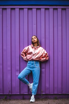 Stylish woman in metallic jacket and jeans posing confidently against a bold purple wall.