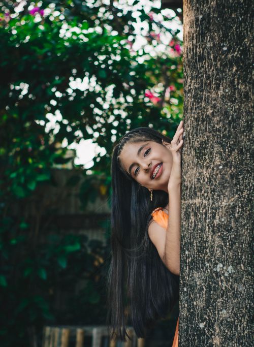 Free Cheerful girl with long hair peering from behind a tree in a nature setting, conveying joy. Stock Photo
