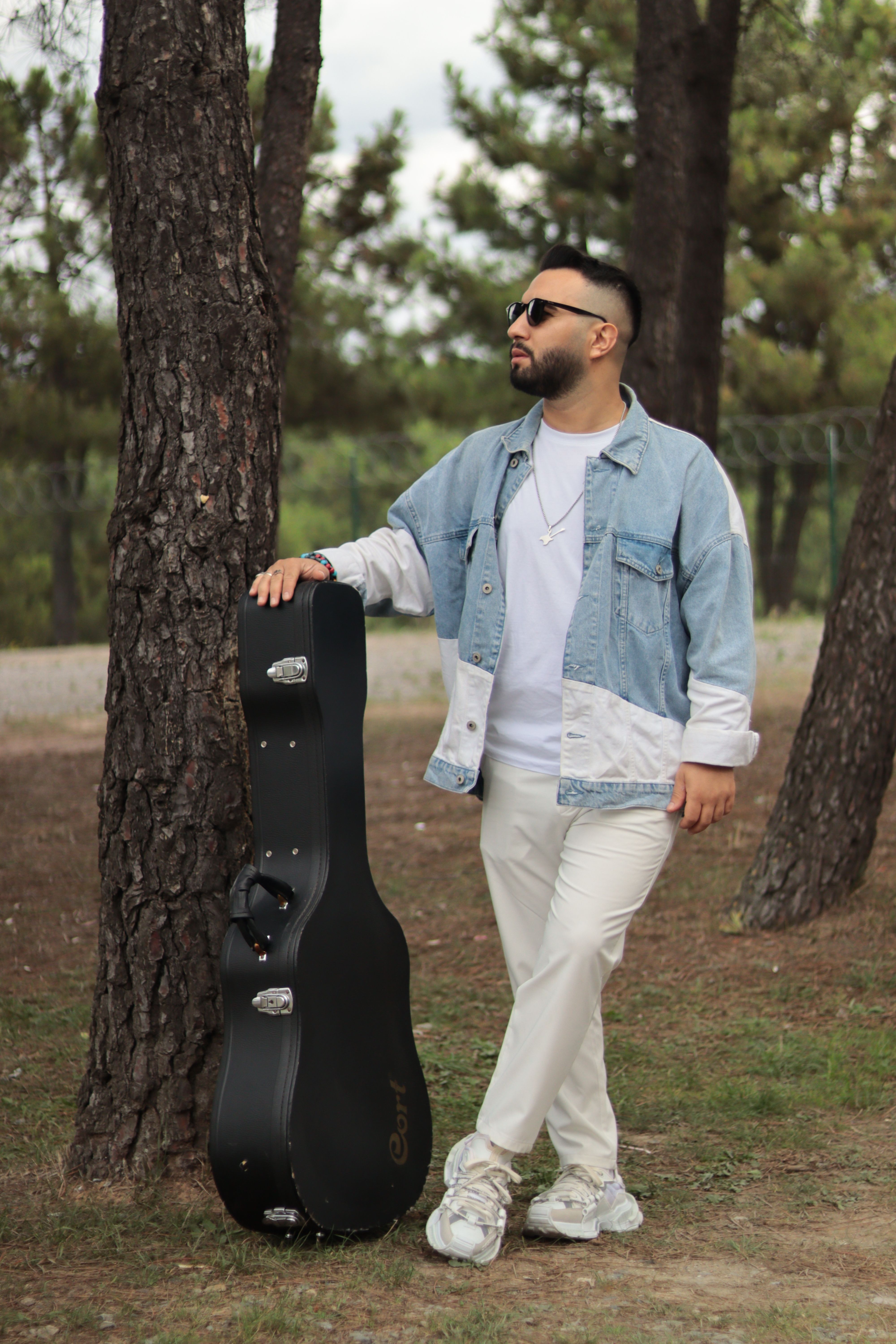 A fashionable man in a denim jacket stands with a guitar case in a pine forest outdoors.