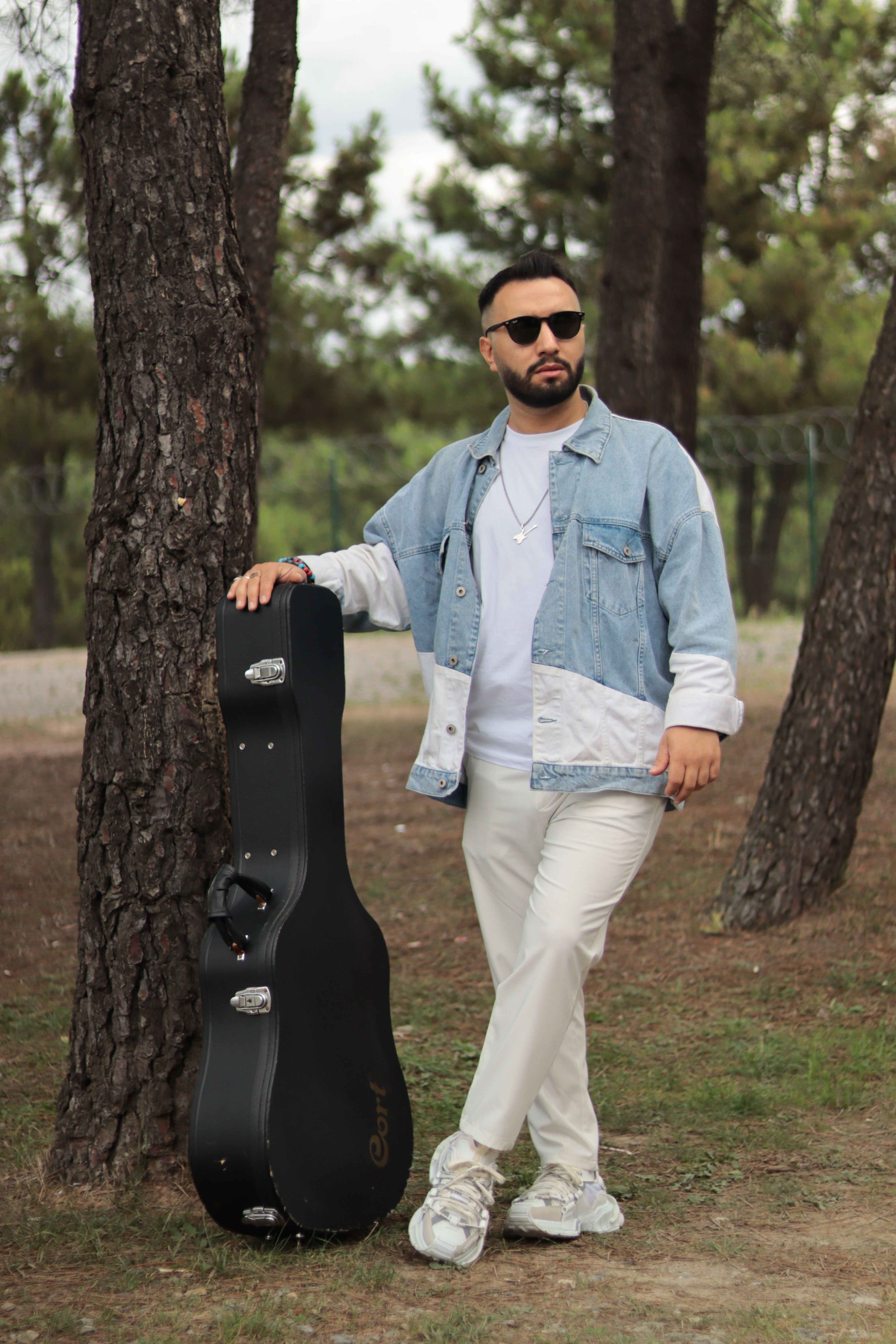 Stylish man in casual outfit leans on guitar case among trees.