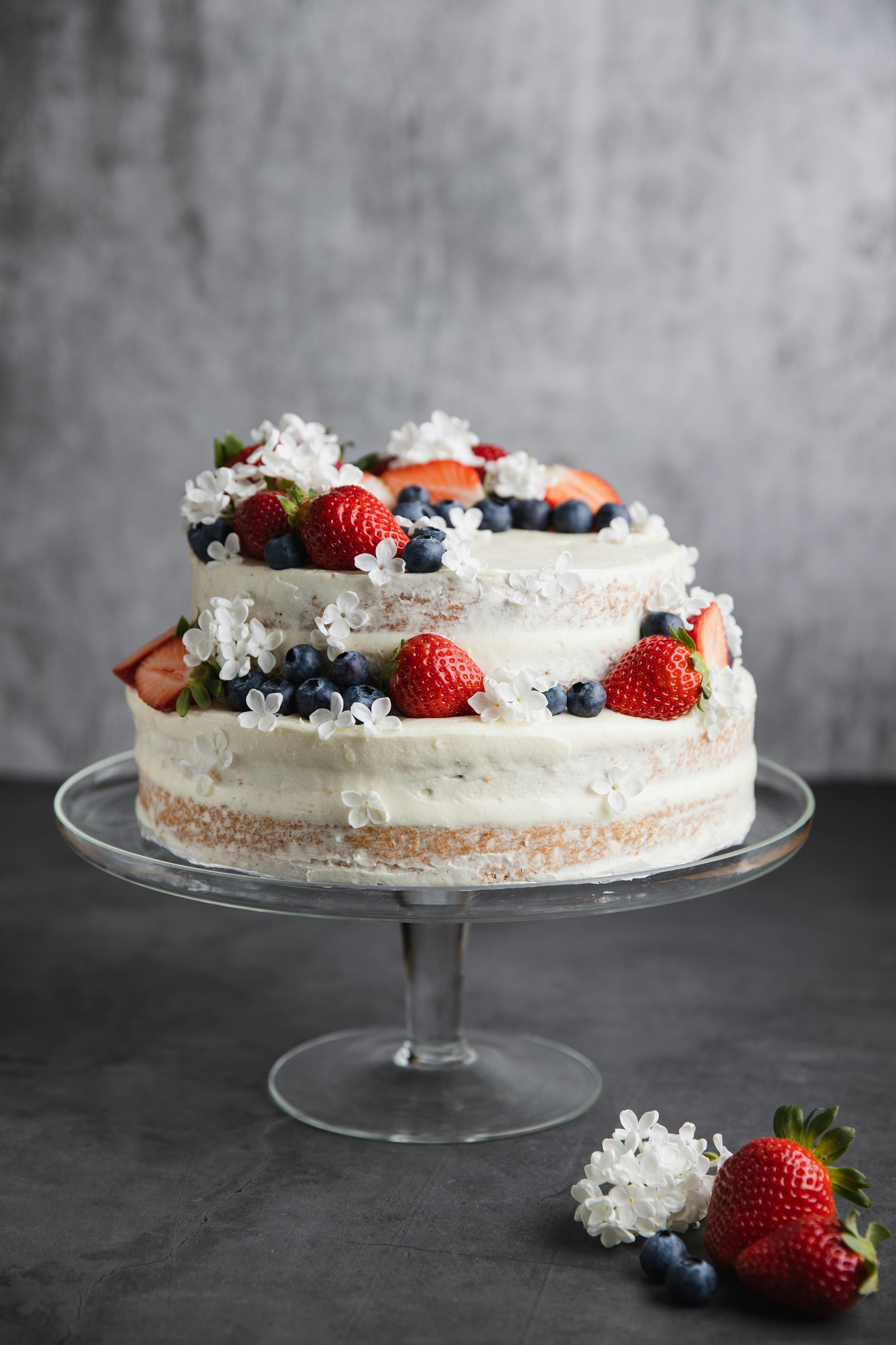 Delicious rustic naked cake topped with fresh strawberries, blueberries, and white flowers on a glass stand.