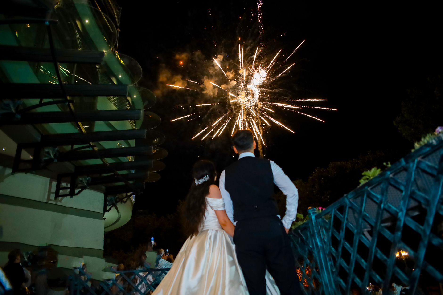 A couple in formal attire watches a dazzling fireworks display at an outdoor event.