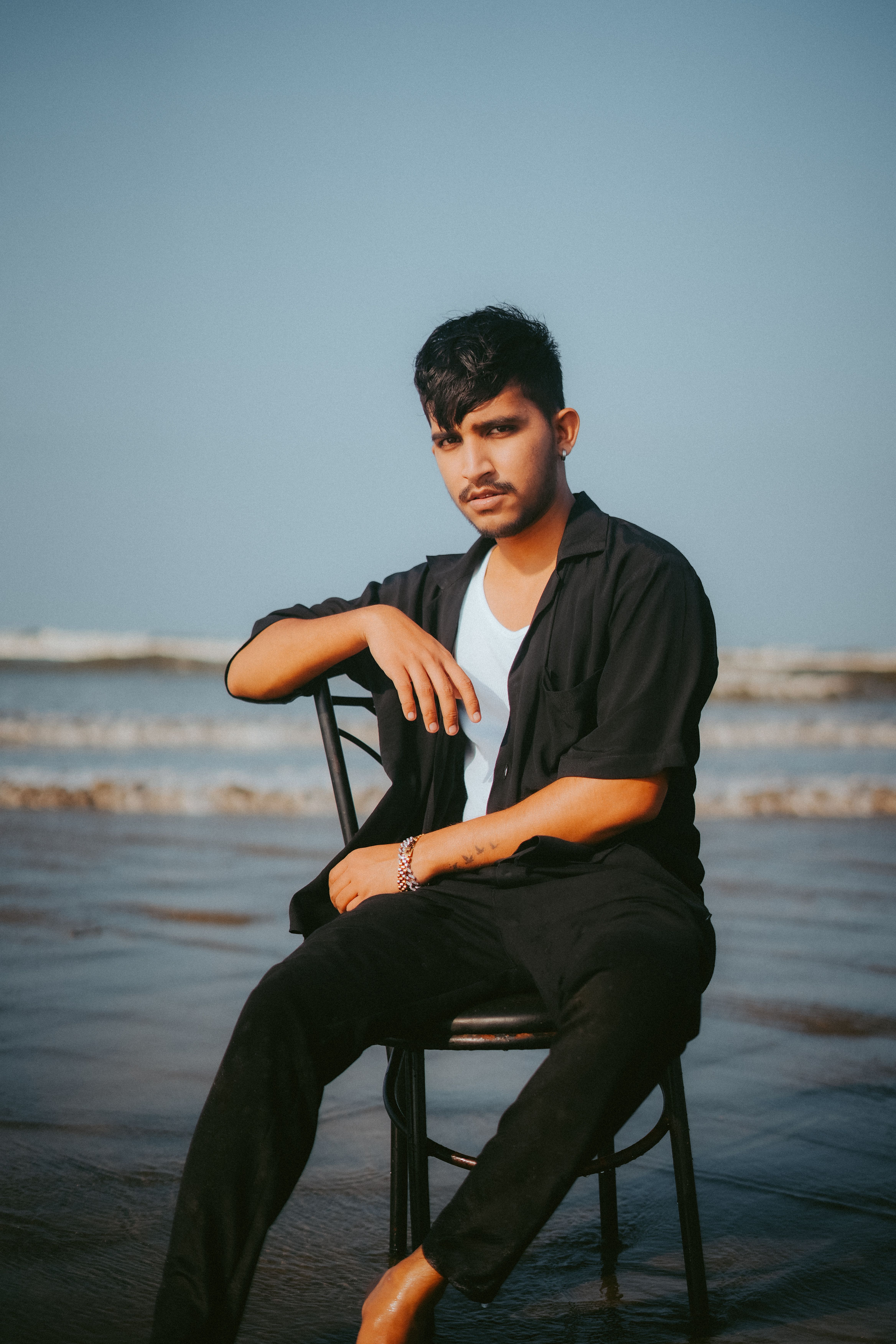 A fashionable man poses on a chair at the beach in Dubai, showcasing modern beach attire.