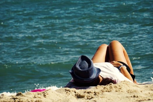 Woman in a hat relaxing on a sandy beach with a view of the ocean under the summer sun.