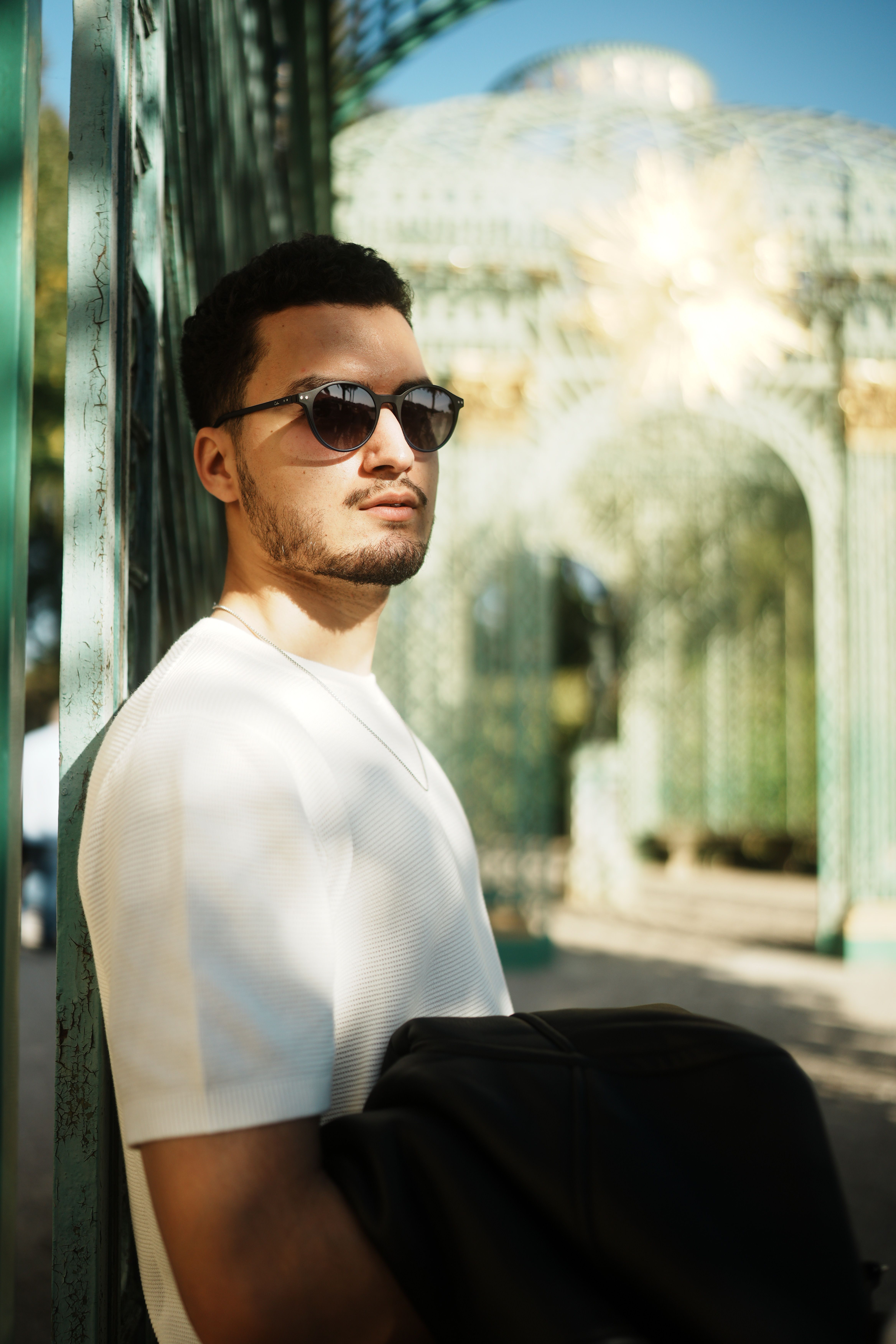 Stylish man wearing sunglasses stands by an ornate iron gazebo in a sunny outdoor setting.