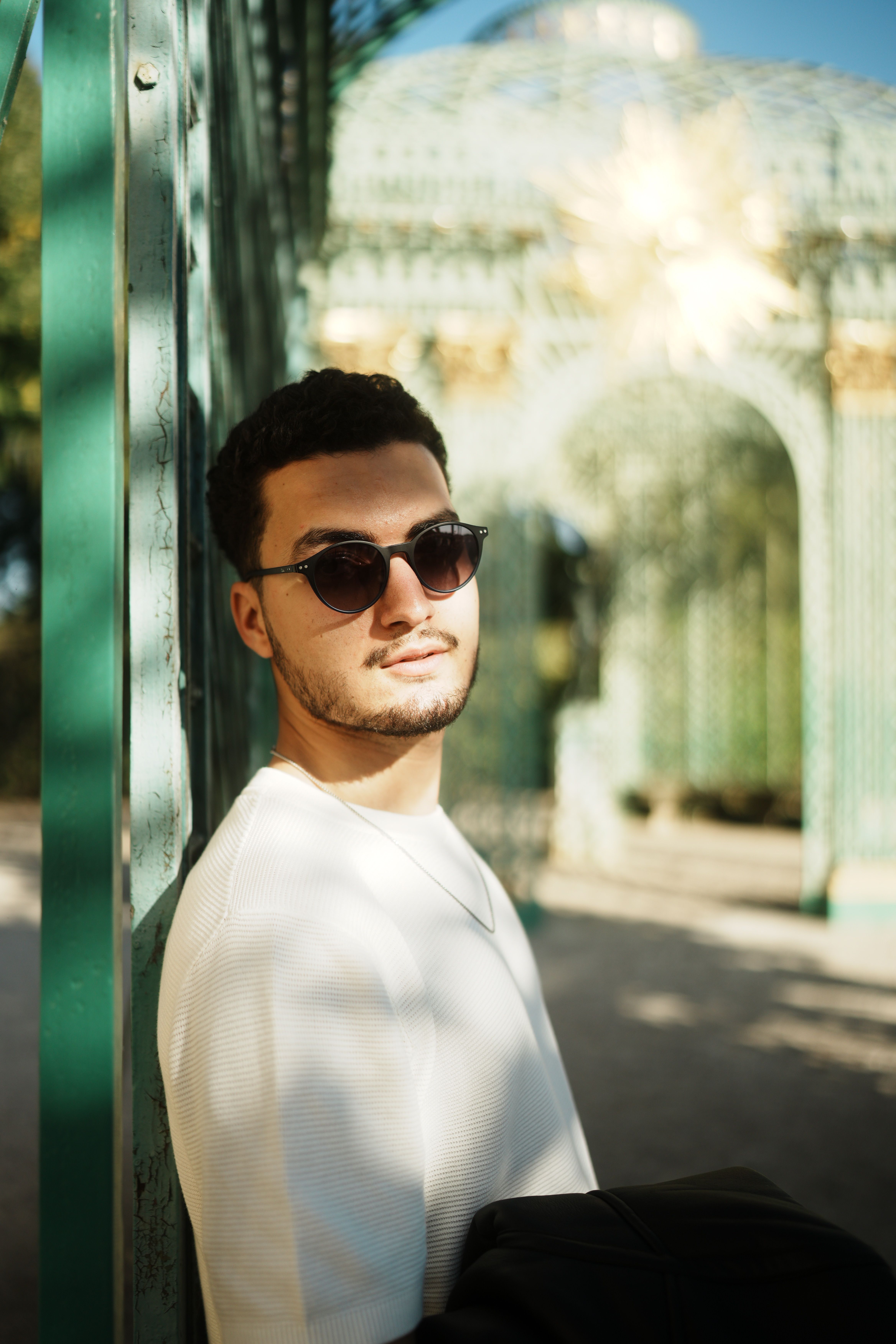 Young man in sunglasses leaning against a green structure outdoors.