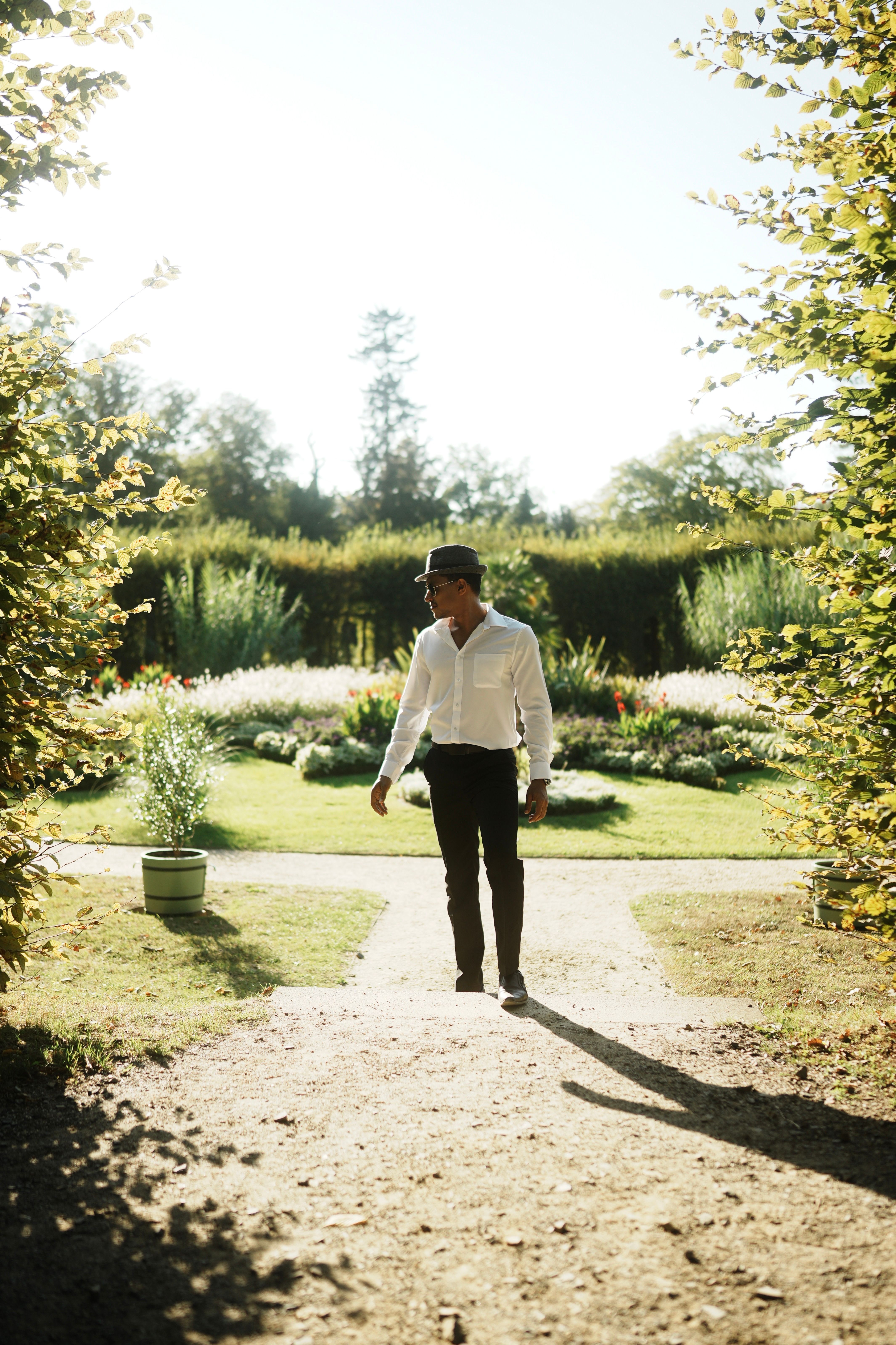 A man in smart casual attire strolls through a sunlit, lush garden path.