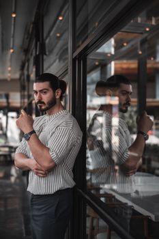 Thoughtful man with a beard stands in an urban cafe, reflected in glass.
