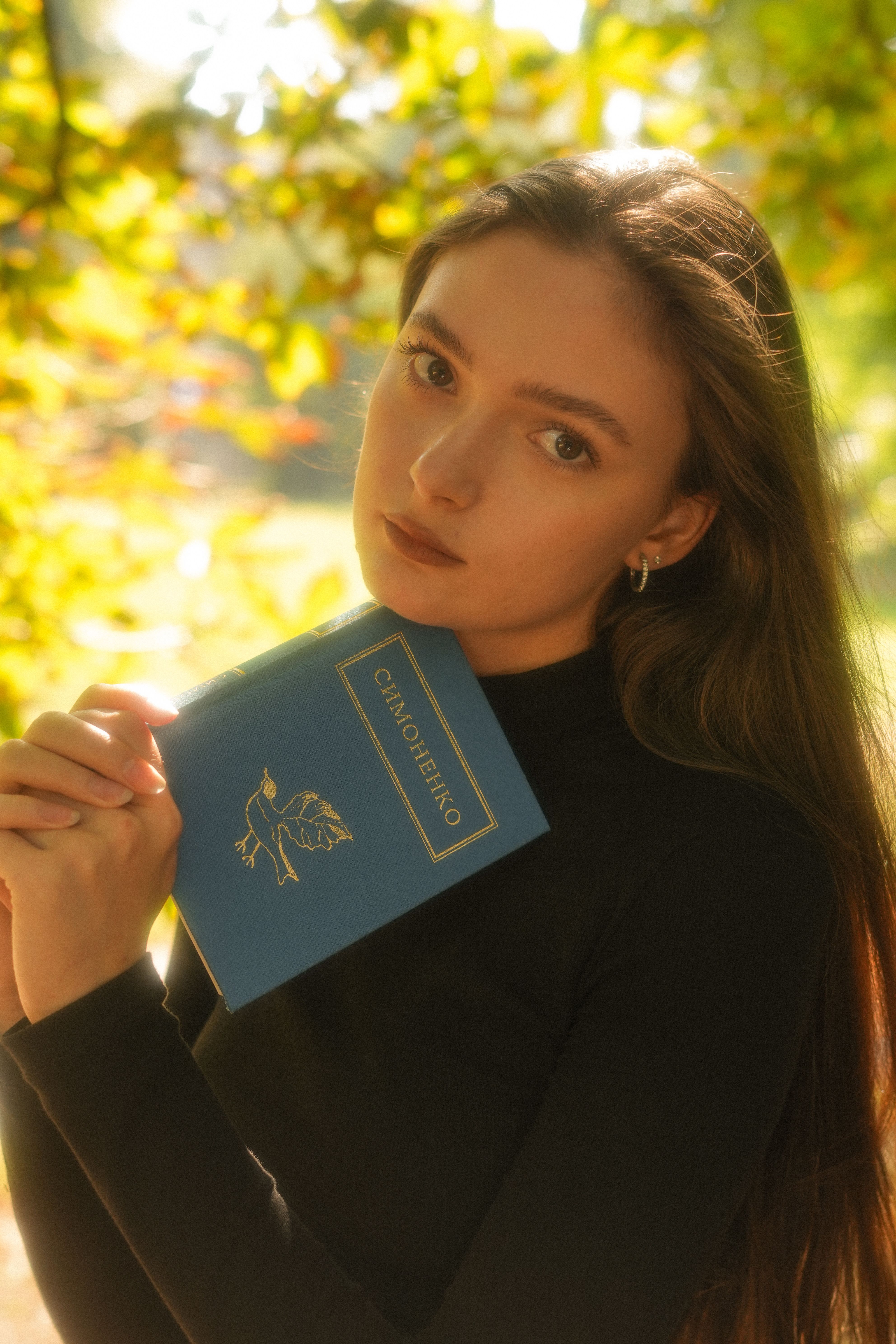 Portrait of a woman holding a blue book in a sunlit outdoor setting with warm tones.