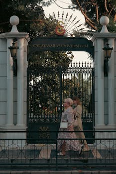 Two elegantly dressed women walk past the ornate gate of Galatasaray University in Istanbul, Turkey.