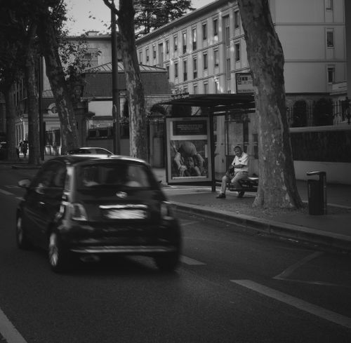 Free Black and white street capture of a moving car and bus stop with a seated person. Stock Photo