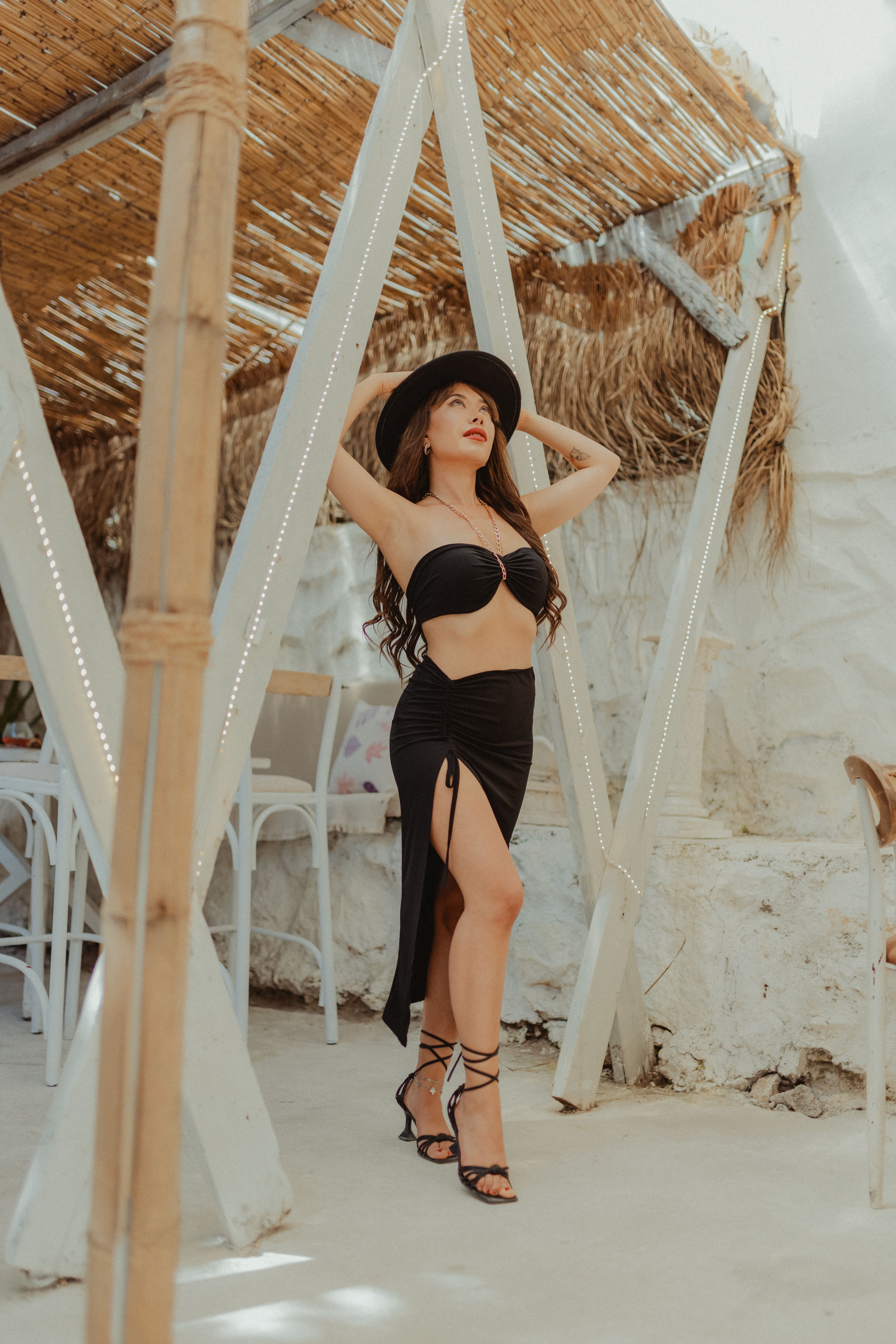 Woman wearing black bikini and hat posing confidently outdoors at a beachside cafe.