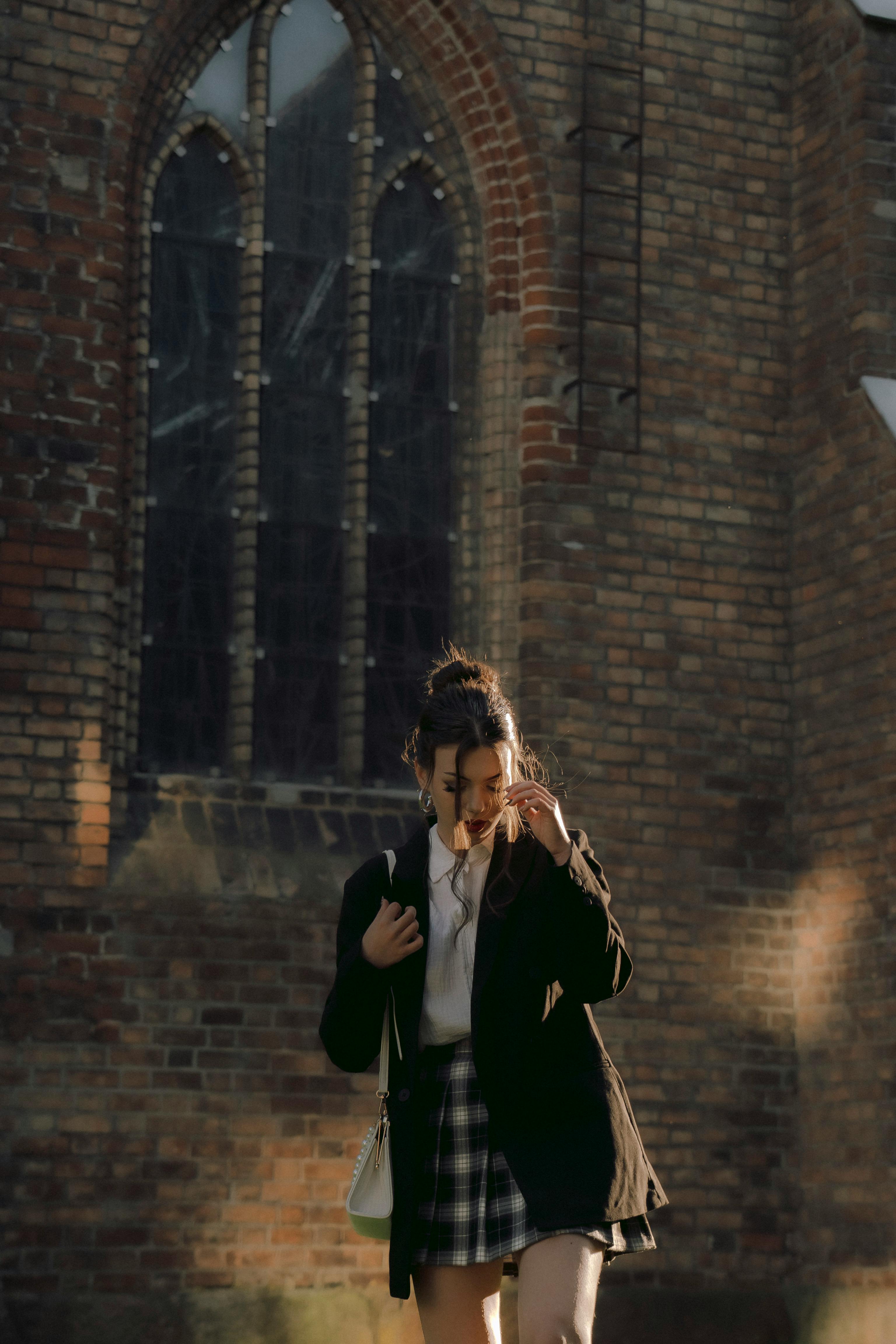 A young woman in a plaid skirt walks by an old brick building on a sunny day.