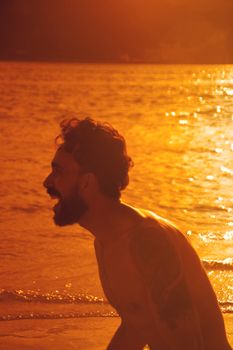 A joyful man with a tattoo laughs by the sea during sunset, embraced by golden light.