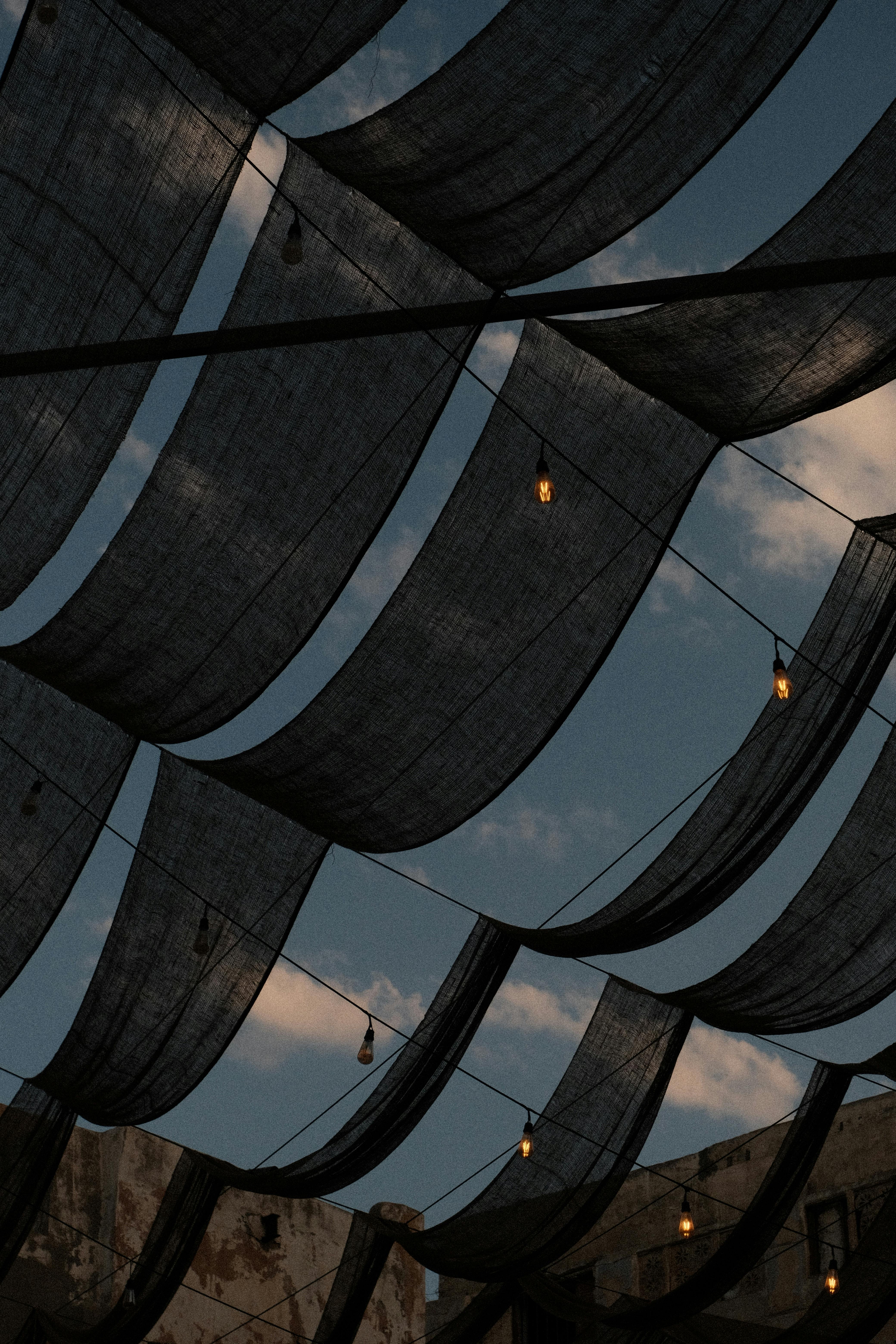 Wire-strung fabric canopies with hanging bulbs against a twilight sky, showcasing Middle Eastern architecture.